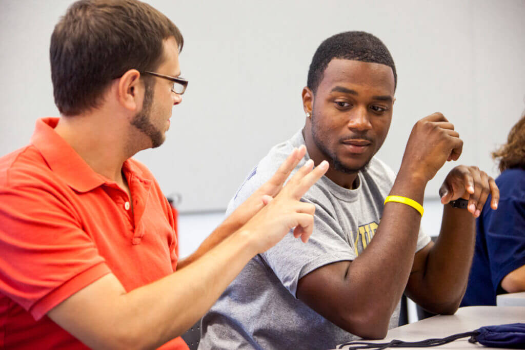 Two men are seated at a table, engaged in conversation. One man, wearing an orange shirt and glasses, is using hand gestures to communicate. The other man, dressed in a gray shirt, is listening attentively, with a thoughtful expression. The background is a neutral-colored wall, suggesting a classroom or meeting setting.