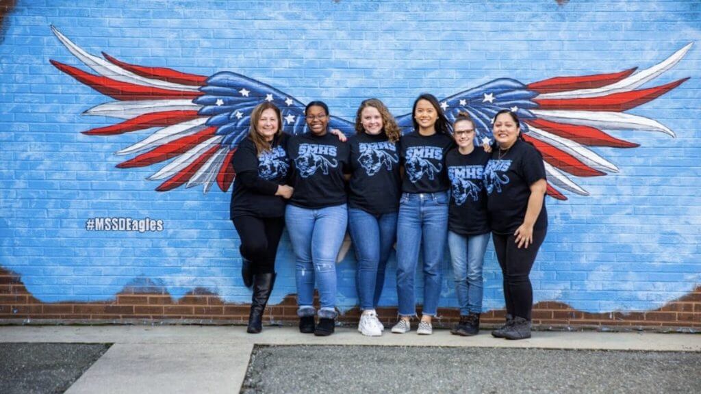 A group of six women standing together in front of a mural featuring large red, white, and blue wings against a blue background. They are all wearing matching black t-shirts with a design on the front and jeans. The women are smiling and posing for the photo.