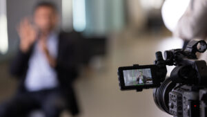 A photo with an in-focus video camera in the foreground, in the bottom right. The out-of-focus background has a seated person signing on the left side.