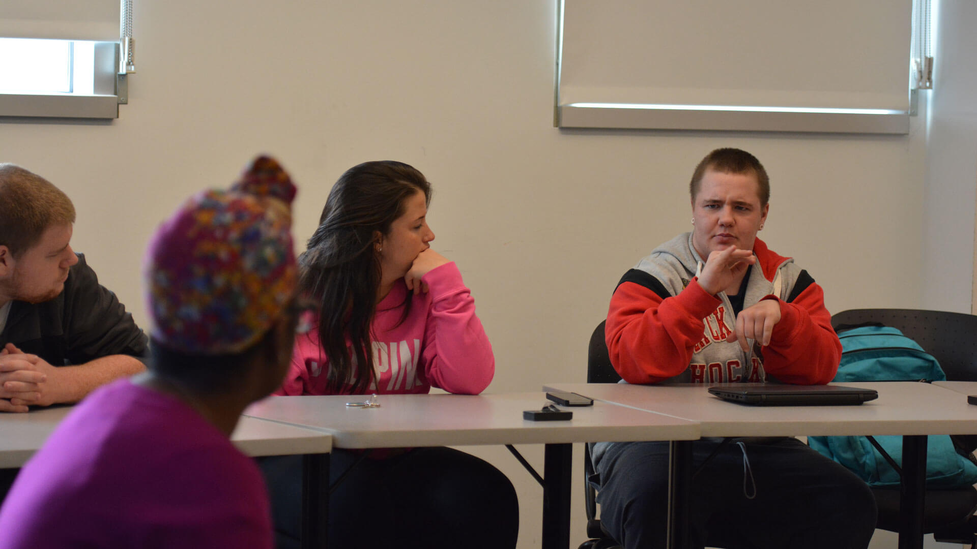 A group of students sitting around a table in a classroom setting, engaged in discussion. One student in the foreground is speaking, while others are listening attentively. The environment is bright, with large windows allowing natural light to enter.