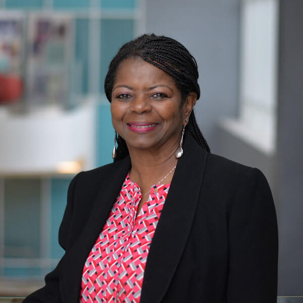 A smiling woman with braided hair wears a black blazer over a red and white patterned blouse. She stands in an indoor setting with blurred backgrounds, conveying a professional and friendly demeanor.