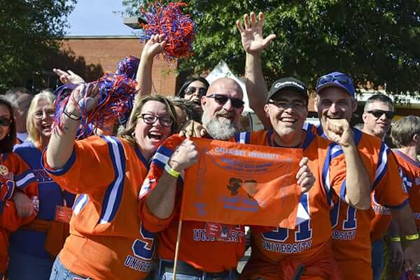 A group of enthusiastic fans wearing orange jerseys and accessories, smiling and celebrating together. They are holding a bright orange sign and cheering, surrounded by trees and a festive atmosphere.