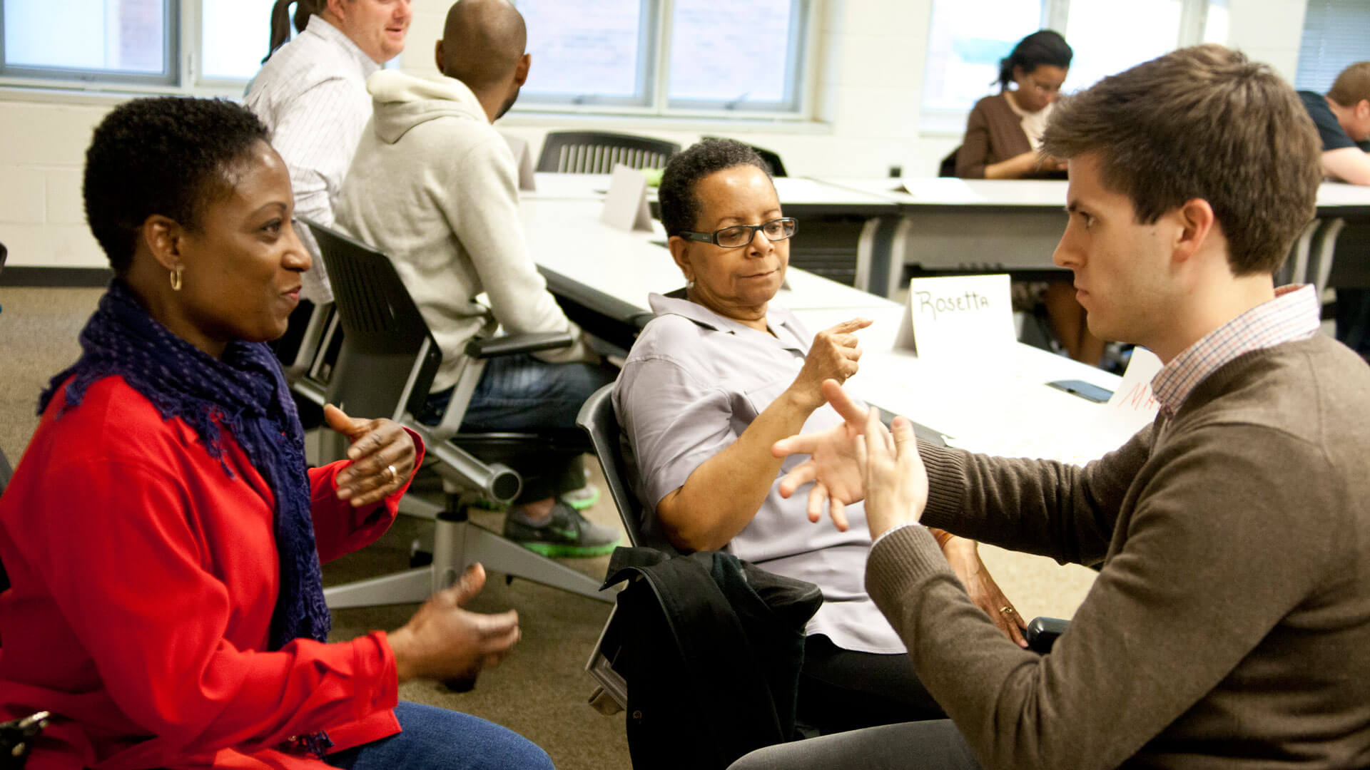 A group of people engaged in conversation in a classroom setting. Two women are seated at the forefront, one wearing a red scarf and the other with glasses, while a man in a brown sweater actively gestures as he speaks with them. In the background, other individuals are seated at tables, some also in conversation.