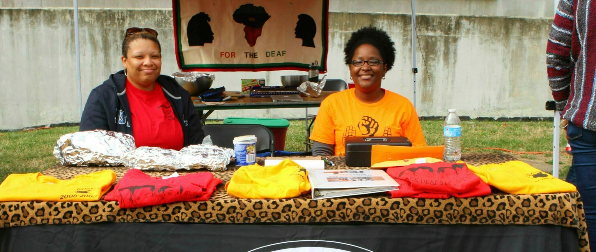 Two women are sitting behind a table at an outdoor event. The table is covered with a leopard print cloth and displays various colored t-shirts (yellow, red) and a few items. Behind them, there is a decorative banner featuring African silhouettes. One woman is wearing a red shirt and the other is in an orange shirt with a logo. There are drinks and food items on the table.