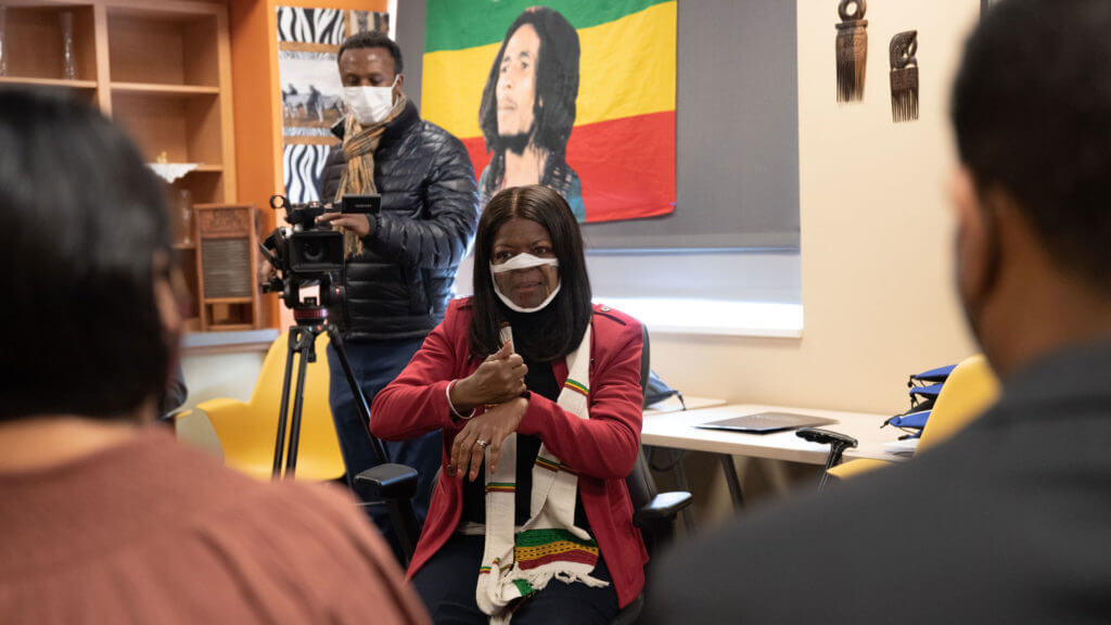A woman wearing a mask sits in a chair, gesturing with her hands while speaking. In the background, a man with a camera stands, and a colorful flag featuring a portrait is visible on the wall. The setting appears to be an indoor space with furniture and decorations.