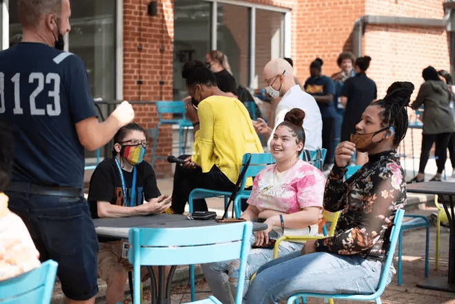 A group of people socializing outdoors at a café, sitting at blue tables and chairs. Some individuals are wearing masks, and there is a mix of casual clothing. A person is standing and speaking to a seated group, which includes two women smiling and engaged in conversation. The setting appears to be a lively and relaxed atmosphere.