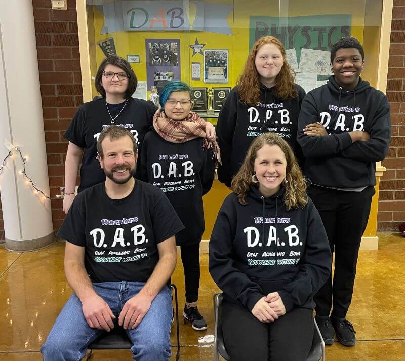 A group of six people posing for a photo in a school setting, all wearing matching black t-shirts that say 