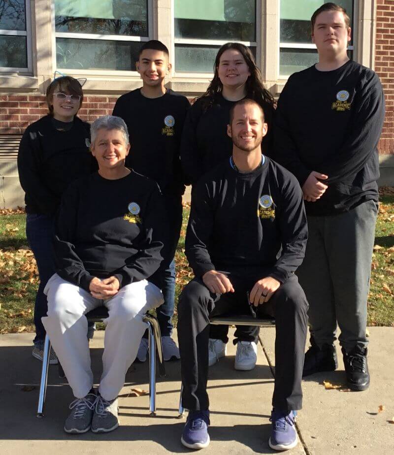 A group of six people poses for a photo outside in front of a brick building. The individuals are wearing matching black sweatshirts with a logo. Two people are seated in front, while four stand behind them. The background features windows and some fallen leaves on the ground.