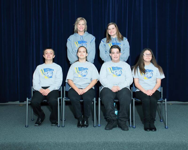 A group of six individuals is seated in front of a dark blue curtain. The front row consists of three people wearing matching gray sweatshirts, while the back row has three individuals, one wearing a lighter gray sweatshirt. They are all smiling and appear to be posing for a photo.