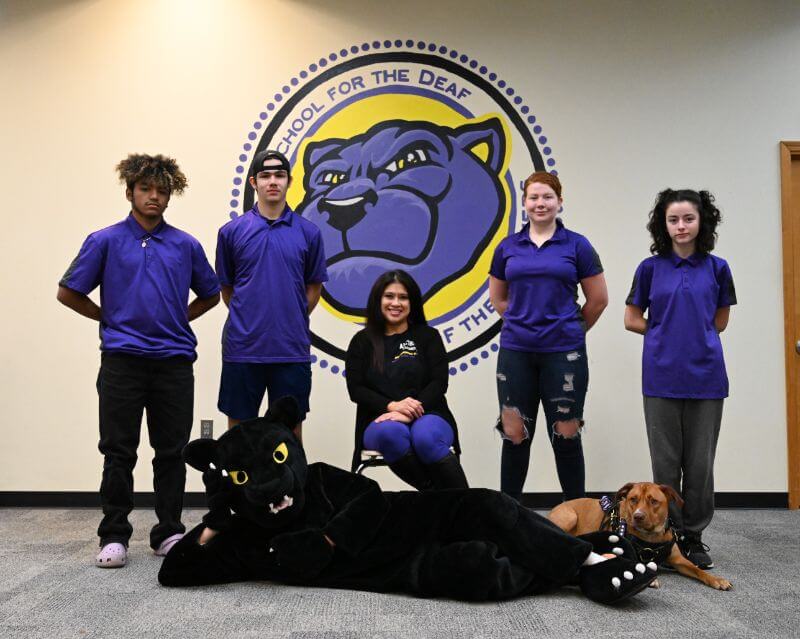 A group of five individuals wearing purple polo shirts pose in front of a large mural of a panther on the wall. One person is seated in the center, while two are standing on each side. A person in a black panther costume lies on the ground, and a dog is resting beside them. The backdrop features the words 
