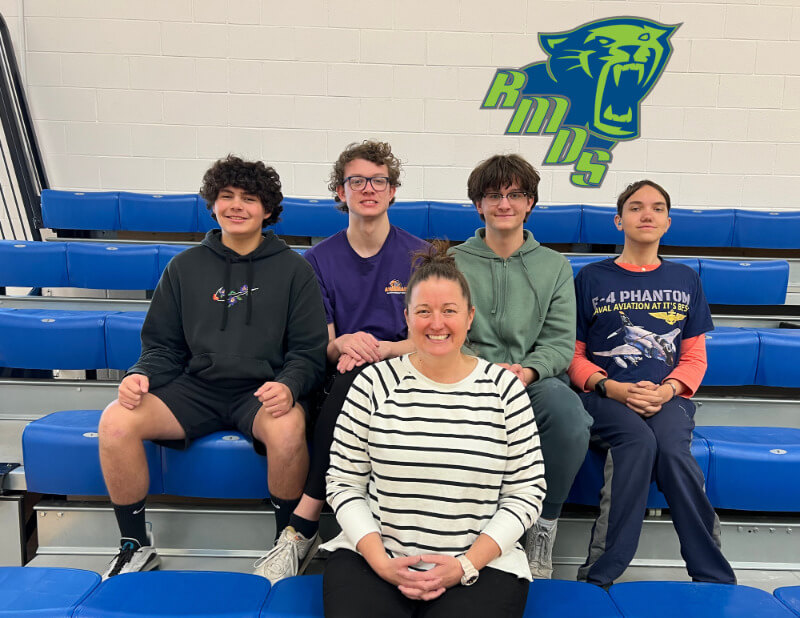 A group of five individuals sitting on blue bleachers in a gymnasium. The front row features a woman in a striped shirt, while four young people are seated behind her. The background includes a logo of a panther and the letters 
