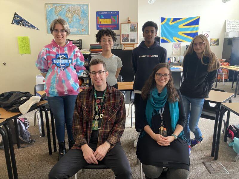 A group of six individuals poses in a classroom setting. Two adults are seated in front, with one man wearing glasses and a plaid shirt, and a woman in a teal scarf. Behind them, four students stand, with a mix of hairstyles and clothing styles, including a colorful hoodie. The background features a world map and a colorful flag. Desks are visible in the classroom.