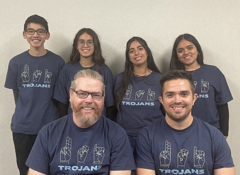 A group of six individuals is posing for a photo, wearing matching dark blue t-shirts that say 