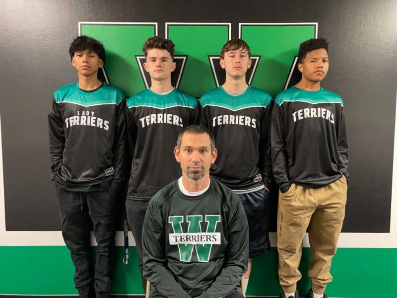 A group of five individuals posing for a photo in a gym or sports setting. Four young athletes are standing at the back, wearing matching black and teal team jerseys with 
