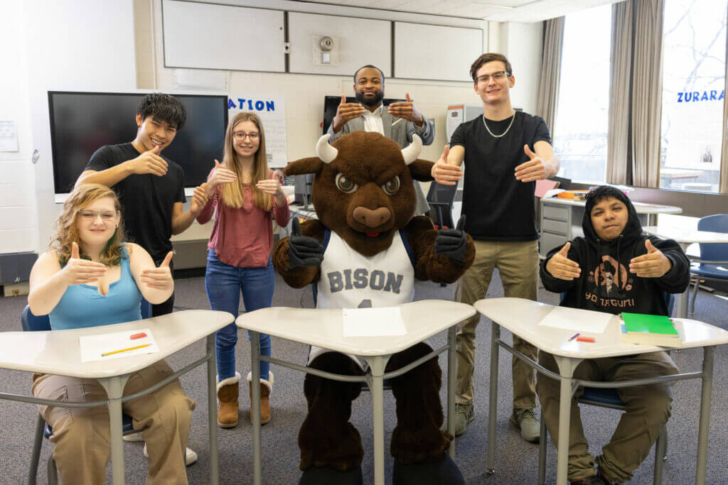 A group of six students and a bison mascot pose together in a classroom setting, all giving thumbs up. The students are seated at desks, with a television screen in the background displaying the word "INFORMATION." The bison mascot, wearing a jersey with the word "BISON," stands at the center of the group.