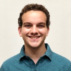 A young man with curly hair is smiling, wearing a teal polo shirt, against a light background.