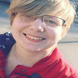 A person with short, light-colored hair is smiling and wearing glasses. They are dressed in a bright red jacket and appear to be outdoors. The background is slightly blurred, focusing on their cheerful expression.