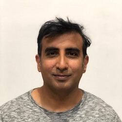 A man with dark hair wearing a gray t-shirt, standing in front of a plain white background, looking directly at the camera.