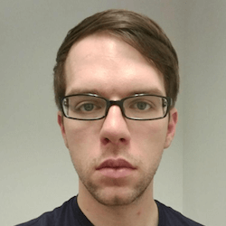A young person with short brown hair and glasses, looking directly at the camera with a neutral expression. They are wearing a dark t-shirt, and the background appears to be a light-colored wall.