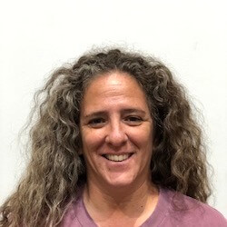 A woman with curly, shoulder-length hair smiling at the camera, wearing a pink shirt, against a plain white background.
