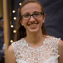 A smiling young woman with long hair, wearing glasses and a white lace top, stands in front of a softly lit background.