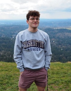 A young person stands outdoors on a grassy hillside, wearing a gray sweatshirt with 