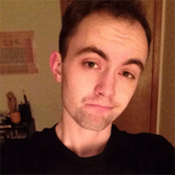 A young man with short hair and a slight smile takes a selfie, wearing a black shirt. The background features soft lighting and a glimpse of a room with various items on a shelf.