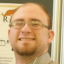 A smiling person with glasses and short hair, wearing a light-colored shirt, standing in front of a display board.