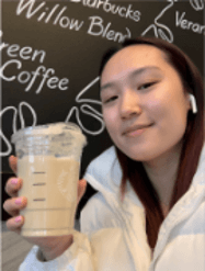A young person smiling and holding a clear cup of iced coffee, wearing a white jacket and earbuds, with a coffee shop menu in the background.