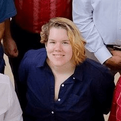 A person with short, wavy blonde hair wearing a navy blue button-up shirt, smiling while seated in a group photo with several other individuals in the background.