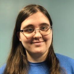 A person with long brown hair and glasses smiles at the camera, wearing a blue shirt against a light blue background.