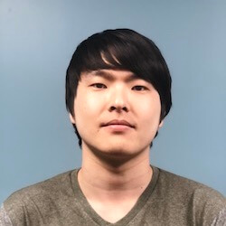 A young man with dark hair and a neutral expression, wearing a gray shirt, is facing the camera against a light blue background.