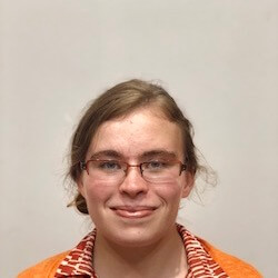 A young person with glasses and light brown hair, wearing an orange jacket over a patterned shirt, smiles at the camera against a plain background.
