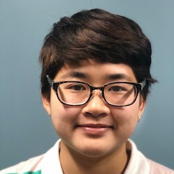 A young person with short dark hair and glasses, smiling in front of a light blue background. They are wearing a collared shirt with colorful stripes.