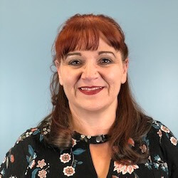A woman with long, reddish-brown hair wearing a floral blouse smiles at the camera against a light blue background.