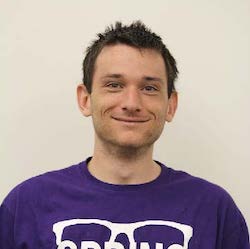 A smiling young man with short, messy dark hair wearing a purple t-shirt with white text. The background is plain and light-colored.