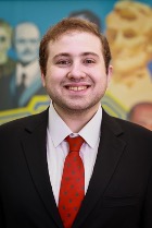 A smiling young man wearing a black suit, white shirt, and a red tie with polka dots, standing in front of a colorful background featuring portraits of historical figures.