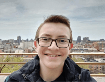 A smiling young person with glasses stands in front of a city skyline, with buildings and a cloudy sky in the background.