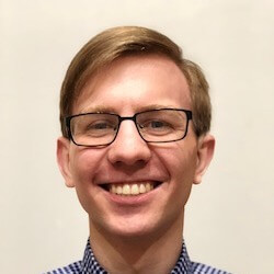 A young man with light brown hair and glasses smiles at the camera, wearing a blue and white checkered shirt. The background is plain and light-colored.