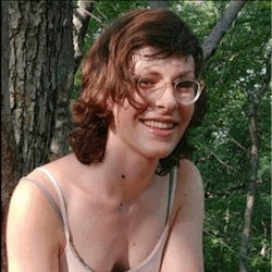 A person with short, wavy brown hair and glasses smiles while sitting outdoors in front of a tree, surrounded by green foliage. They are wearing a light-colored sleeveless top.