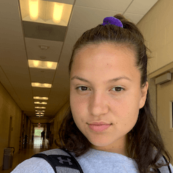 A young person with dark hair pulled back in a ponytail, wearing a gray sweatshirt, stands in a brightly lit hallway with doors and ceiling lights visible in the background.