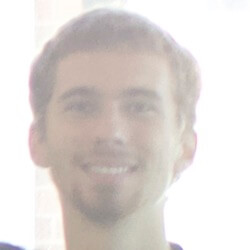 A smiling young man with light brown hair and a goatee, standing in front of a bright background that partially obscures his features.