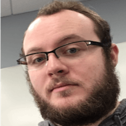 A close-up portrait of a person with glasses and a beard, looking directly at the camera. The background is blurred and shows a light-colored wall. The person is wearing a gray hoodie.