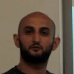 A man with a bald head and a beard is looking towards the camera. He appears to be in an indoor setting with a light-colored wall in the background.