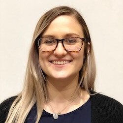 A smiling woman with long, straight blonde hair, wearing glasses and a dark sweater, poses against a light background.
