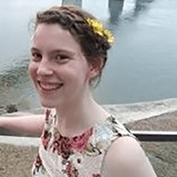 A young woman with a braided hairstyle adorned with a yellow flower, smiling while standing near a body of water. She is wearing a floral dress and is positioned in a natural outdoor setting.