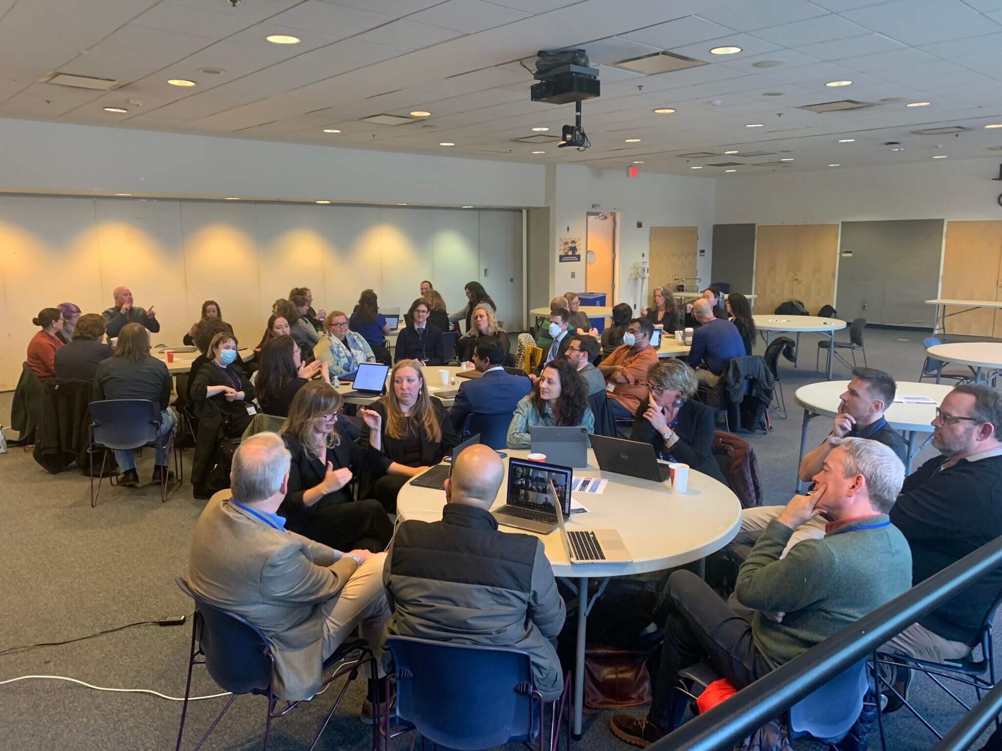 A group of people seated at round tables in a conference room, engaged in discussions. The room is well-lit, with several individuals using laptops. There are diverse attendees, and the atmosphere appears collaborative and interactive.