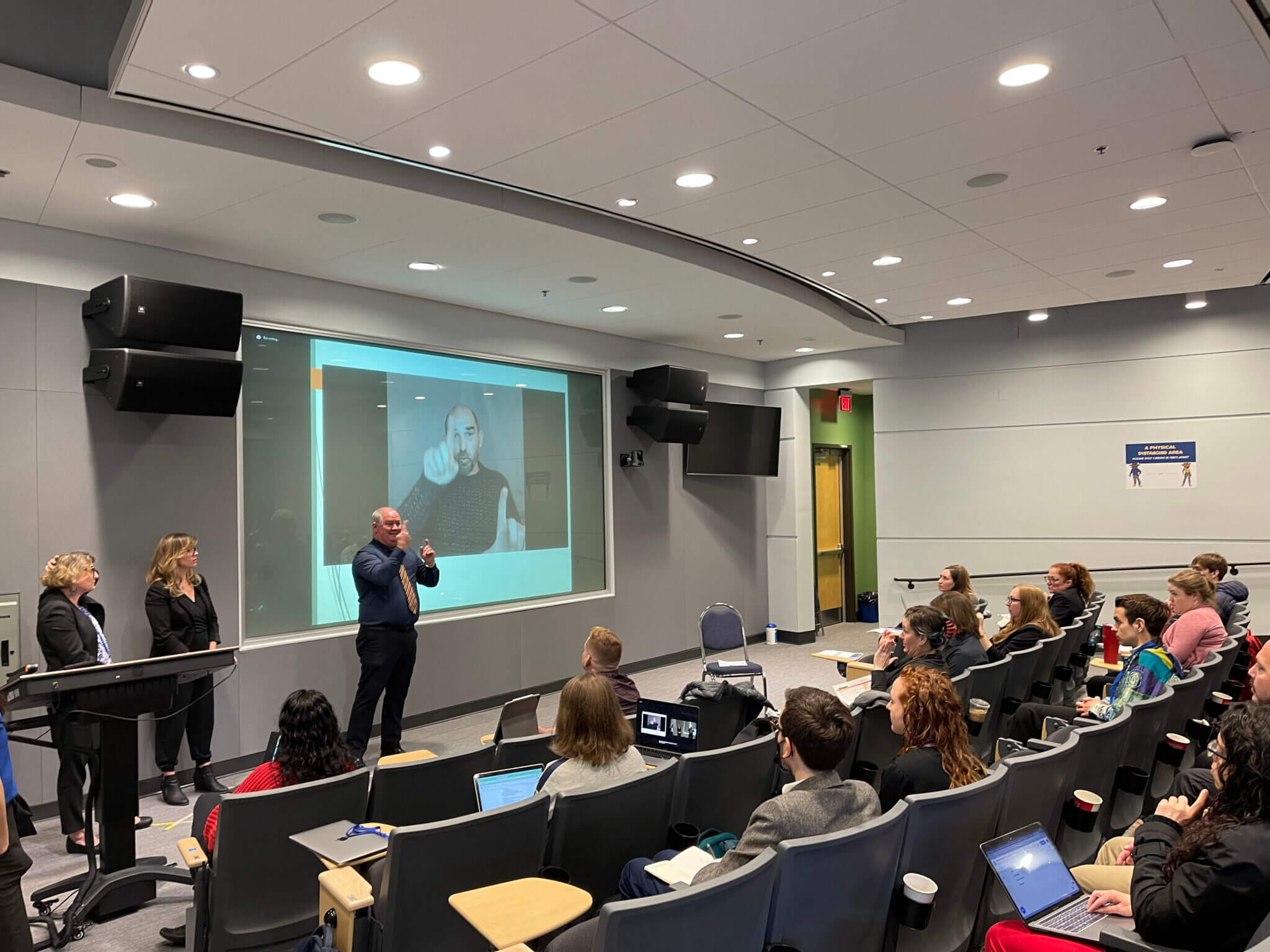 A presentation is taking place in a classroom setting. A speaker stands at the front, gesturing while two women flank him. Behind them, a large screen displays an image. An audience of students sits at desks, some with laptops open, attentively watching the presentation. The room is equipped with audio-visual technology.