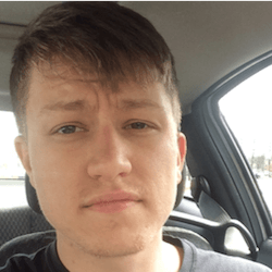 A close-up selfie of a young person with short, light brown hair and a slight stubble, sitting inside a car. The background shows the interior of the vehicle and a glimpse of the outside through the window.