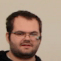 A man with short dark hair and glasses, wearing a black hoodie, appears to be speaking or listening in a light-colored room.